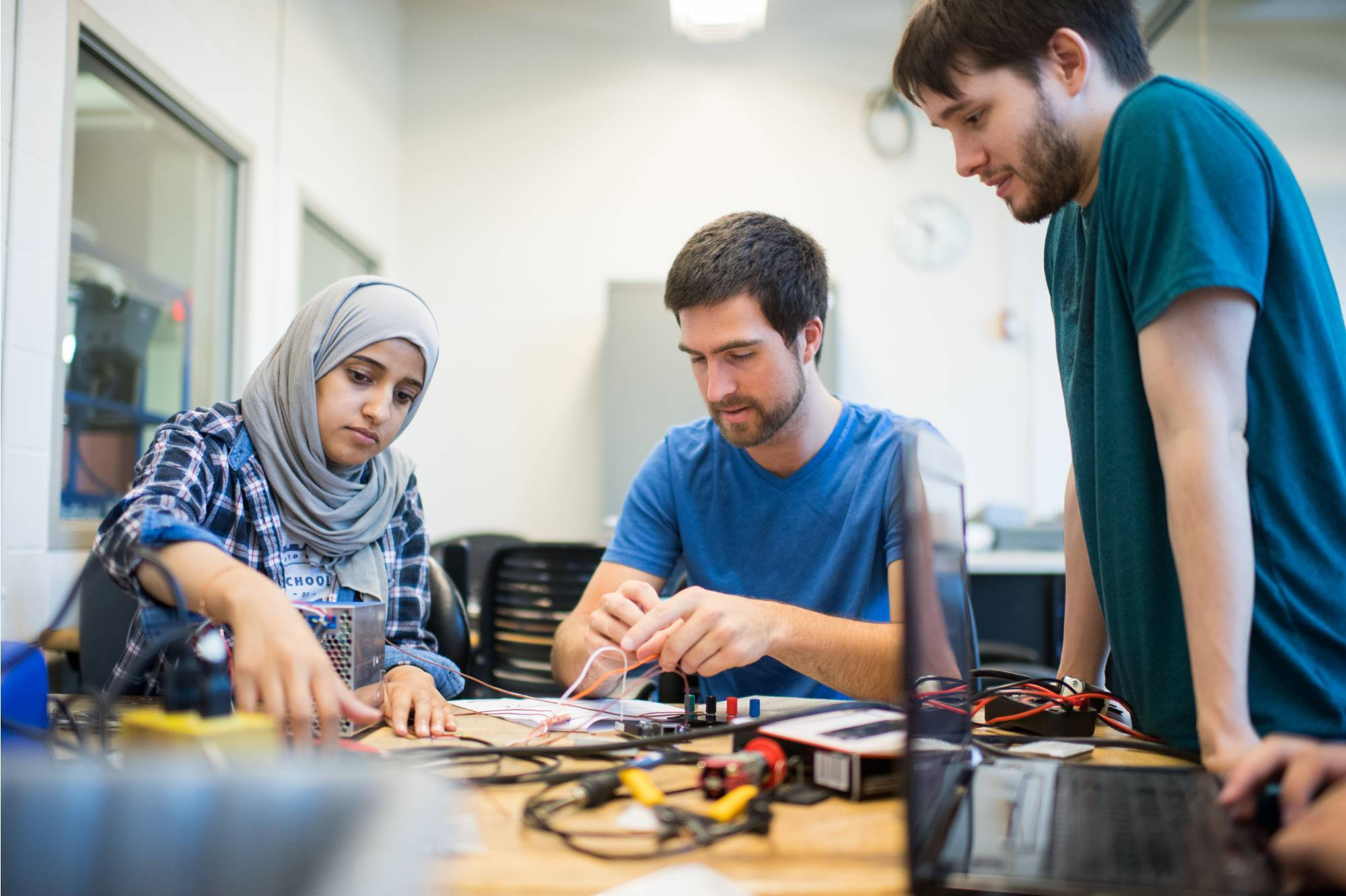 Three students collaborate on an electronics project in a bright lab, focusing on circuit components. The atmosphere is focused and collaborative.